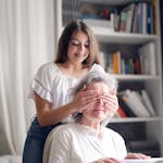 A joyful moment as a young woman playfully surprises her grandmother while reading indoors by covering her eyes.