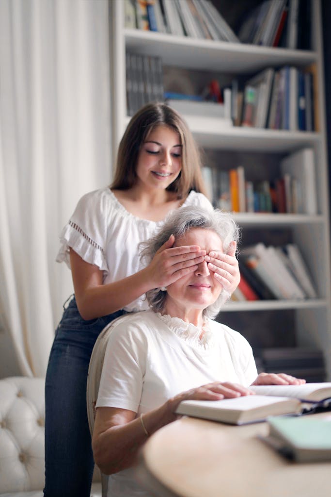 A joyful moment as a young woman playfully surprises her grandmother while reading indoors by covering her eyes.